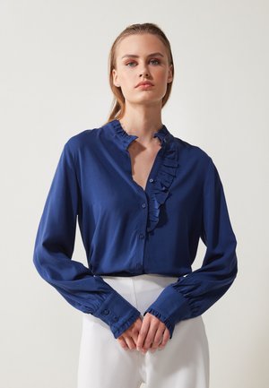 Blå bluse med volangkrave og frontplacket, med lange ærmer og knaplukning ved håndleddene. Fremstillet af et glat stof med en blank overflade.