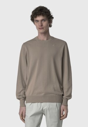 Giovane uomo con capelli ricci che indossa un maglione beige girocollo e pantaloni chiari, in piedi contro uno sfondo semplice.