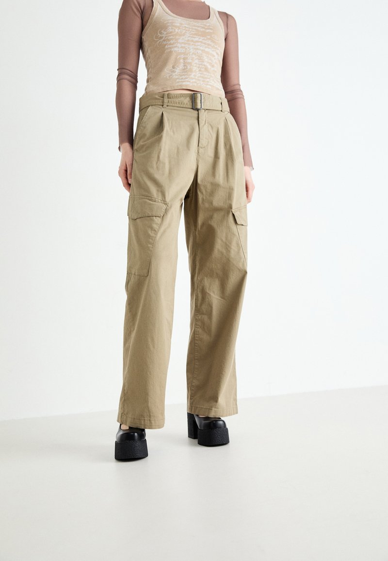 Khakifarbene Cargo-Hosen mit Falten, weitem Bein und zwei seitlichen Taschen. Getragen mit einem hellbraunen Tank-Top und klobigen schwarzen Plateauschuhen.