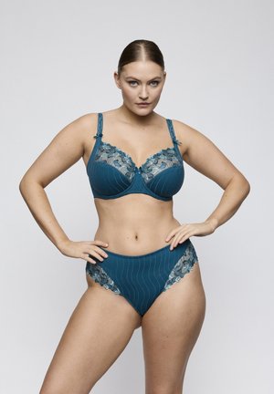 Vrouw die een teal gekleurd kanten lingerieset met bloemmotief draagt, staand met handen in de zij tegen een effen lichte achtergrond.