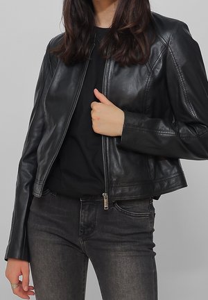 Persona che indossa una giacca di pelle nera sopra una maglietta nera, abbinata a jeans grigio scuro, mentre tiene la zip della giacca con una mano.