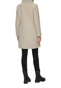 Beige wollen blend jas met een hoge kraag, gestroomlijnde silhouette en lange mouwen, gecombineerd met zwarte aangesloten broek en zwarte veterschoenen.