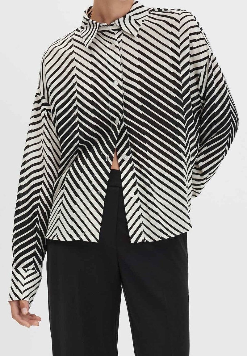 Personne portant une chemise rayée noire et blanche avec des lignes diagonales et un pantalon noir uni sur un fond uni.