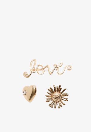 Three gold accessories: a cursive "love" script, a glossy heart with a rhinestone, and a daisy with textured petals.
