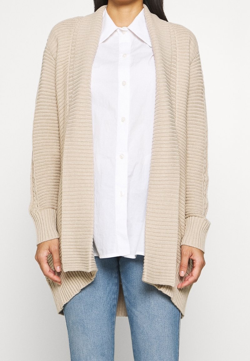 Femme portant un cardigan beige à côtes ouvert sur le devant, par-dessus une chemise blanche à boutons, et un jean bleu clair, debout devant un arrière-plan uni.