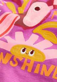 Tissu rose vif avec un tournesol souriant brodé, un oiseau et le mot partiel "SUNSHINE" en lettres orange.