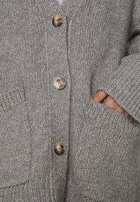 Grå stickad kofta med v-ringning, fyra stora knappar och ribbade ärmslut. Har två framfickor för ökad funktionalitet.