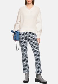 Cremefarbener Strickpullover mit V-Ausschnitt, kombiniert mit marineblauen gemusterten Hosen. Graue Stiefeletten und eine blaue Umhängetasche vervollständigen das Outfit.