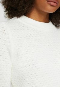 Pull en tricot blanc avec un motif texturé et un col rond à bord côtes. La vue rapprochée met en avant le design respirant du tissu.