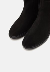 Paire de bottines en daim noir avec bouts arrondis et semelles noires sur fond blanc.