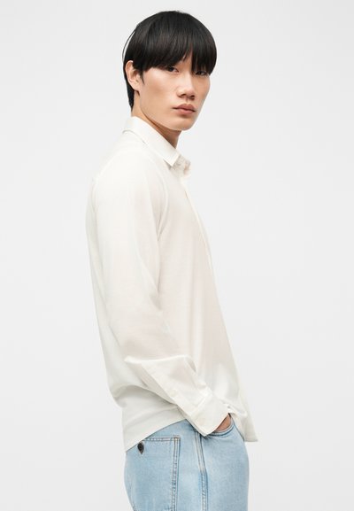 Νέος άντρας με μαύρα μαλλιά, φοράει λευκό πουκάμισο με μακριά μανίκια και ανοιχτό μπλε τζιν, στέκεται πλευρικά με το ένα χέρι στην τσέπη.