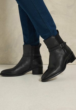 Personne portant un pantalon bleu foncé et des bottines en cuir noir avec des fermetures éclair latérales et des petits talons carrés, debout sur un sol clair.