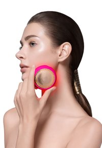 Masseur facial rose avec une surface circulaire texturée dorée, tenu contre la mâchoire, brillant d'un rouge éclatant, sur un modèle ayant une peau lisse.