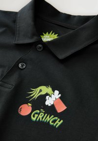 Zwarte polo met een kleurrafische afbeelding van de hand van de Grinch die een ornament vasthoudt, met een groene label waarop "The Grinch" staat.