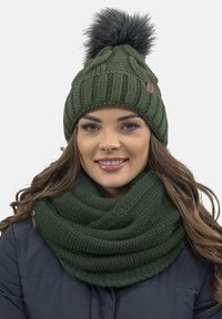 Gorro de punto en verde oscuro con pompón de piel sintética y bufanda tipo cowl a juego, que muestra una textura acanalada y un diseño elegante.