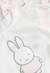 Biały top z tkaniny z szarym, haftowanym wzorem królika. Posiada falbany na ramionach oraz zapięcia na napy, co ułatwia zakładanie.