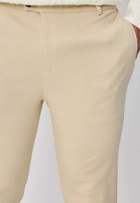 Pantalon beige en tissu lisse ; coupe ajustée avec poches avant et détails de coutures discrètes. Aucun motif ou élément visible.