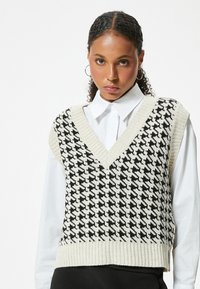 Mustekki harmaavalkoinen houndstooth-kuvioitu neuleliivi V-painatuksella ja ribbaamalla, valkoisen paidan päällä. Sileä pinta ja keskikokoinen istuvuus.