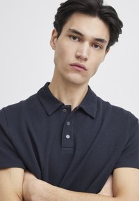 Marineblå polo-skjorte med teksturert stoff, korte ermer og en tre-knapps knapplist. Kragen er flat, og det overordnede designet er tettsittende.