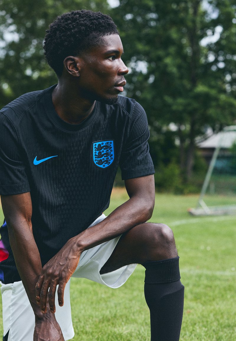 Black Nike football jersey with blue accents and crest, textured fabric, paired with white shorts and black knee-high socks on grass field.