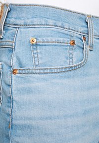 Un bolsillo de jeans de denim azul claro con remaches de cobre y costuras naranjas sobre un fondo blanco.