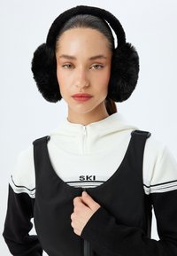 Zwarte faux fur oordoppen met een zachte textuur, gedragen over een witte hoodie met rits en een zwart ski-logo, en een zwarte overall.