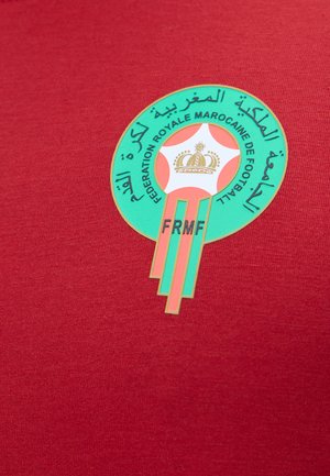 Embleem van de Koninklijke Marokkaanse Voetbalfederatie met een kroon in een ster, Arabische en Franse tekst, op een rode stoffen achtergrond.