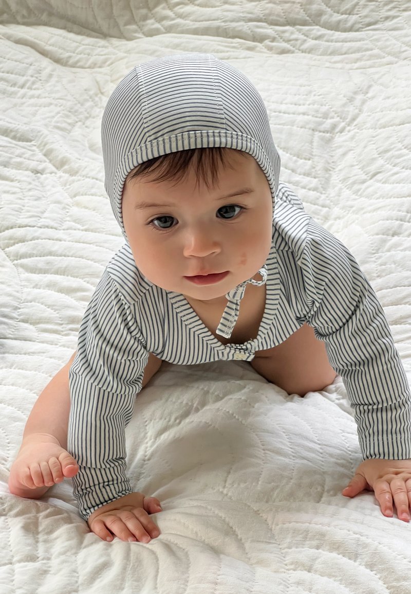 Gestreifter Baby-Body mit passendem Hut, ausgestattet mit einem Druckverschluss und langen Ärmeln, aus weichem Stoff, auf einem gesteppten weißen Hintergrund.