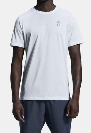 Mandlig model iført en ensfarvet hvid kortærmet sportst-shirt med et lille logo på venstre bryst, kombineret med mørke shorts.
