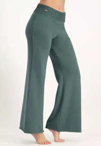 Nainen, joka käyttää teal-värisiä leveälahkeisia housuja, joissa on joustava vyötärö, seisoo varpaillaan yksinkertaista vaaleaa taustaa vasten.