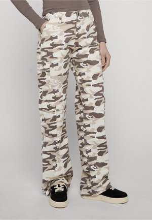 Személy bézs és barna terepmintás cargó nadrágban, fekete, fehér talpú sportcipőben, egyszínű szürke háttér előtt.