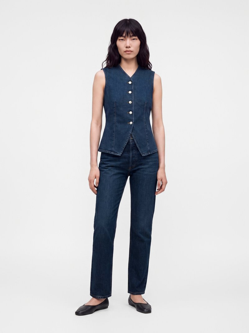 Femme portant un gilet en jean bleu foncé sans manches avec des boutons et un jean droit assorti, debout, les bras détendus le long du corps.