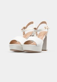 Sandalias blancas de tacón alto con punta descubierta, tira en el tobillo y un tacón en bloque. Material suave con un acabado brillante y una base de plataforma redonda.