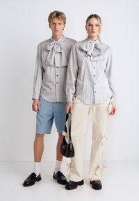 Δύο μοντέλα φορούν ταιριαστές γκρι ριγέ πουκάμισα με λεπτομέρειες φιόγκου, τα οποία συνδυάζονται με ανοιχτό μπλε τζιν σορτς και κρεμ παντελόνια με κορδέλες.