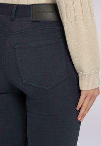 Donkerblauwe jeans met een gladde textuur, met een achterzak in een diamantvorm. Een zwart leren merketiket op de tailleband.