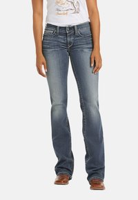 Jeans de mezclilla azul con diseño de pierna acampanada, con costuras en contraste y estilo de cinco bolsillos. Llevados con una camiseta gráfica blanca y botas marrones.