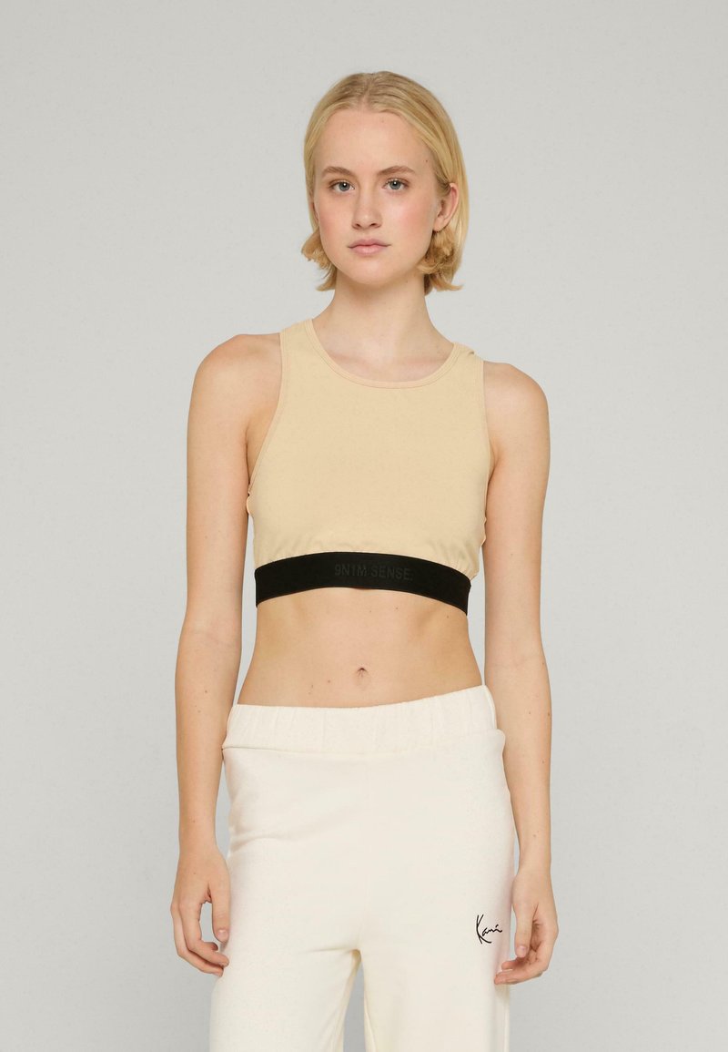 Top scurt bej cu un band elastic negru ce conține text, asociat cu pantaloni de trening crema cu un logo subtil. Textură netedă.