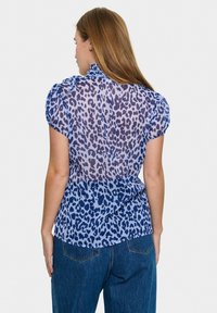 Blusa trasparente blu con design a stampa leopardo. Presenta maniche corte a sbuffo e un colletto alto. La parte posteriore ha una finitura liscia e un leggero arriccio.