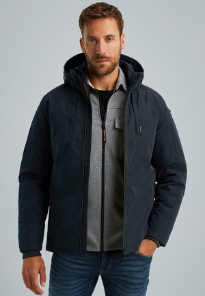 Veste à capuche bleu marine foncé avec une finition texturée, dotée d'une couche intérieure grise et d'une fermeture éclair à l'avant, accentuée par des accessoires jaunes.