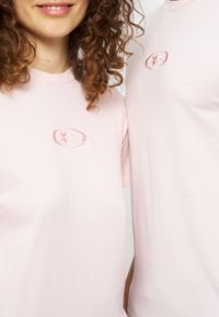 Δύο άτομα φορούν ανοιχτό ροζ T-shirts με μικρά κυκλικά λογότυπα αστεριών στην αριστερή πλευρά του στήθους, εμφανίζονται από τους ώμους έως τη μέση του κορμού.