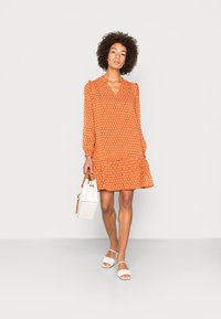 Vestido laranja de bolinhas com mangas longas e bainha franzida, combinado com sandálias brancas e uma mala branca estruturada.