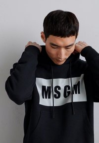 Schwarzer Kapuzenpullover mit einem großen weißen rechteckigen Feld und schwarzem "MSGM"-Text. Glatter Stoff mit einer Fronttasche und Kordelzügen.