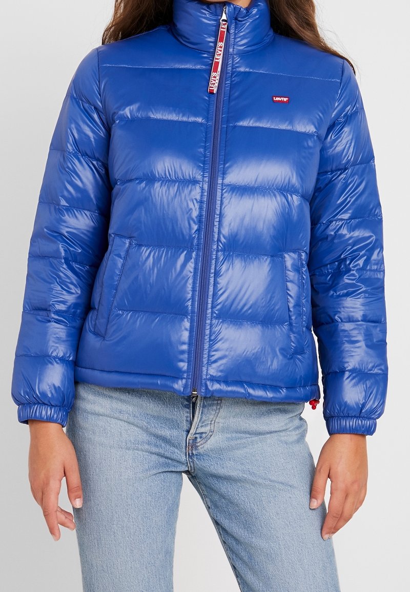 Doudoune bleue Levi's fermée par une fermeture éclair, portée avec un jean bleu clair, montrant un motif matelassé et le logo Levi's sur la poitrine et le curseur de la fermeture éclair.