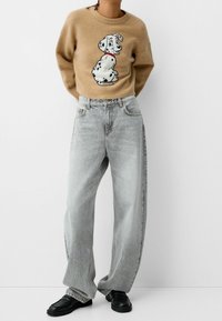 Beiger Pullover mit einem Dalmatiner-Graphic, kombiniert mit verblassten grauen weit geschnittenen Jeans. Das Model trägt schwarze Slipper.