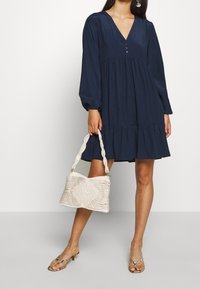 Mörkblå klänning med V-ringning, knapppropp och en tippad fåll. Kombination med en vit vävd handväska och mönstrade sandaler.