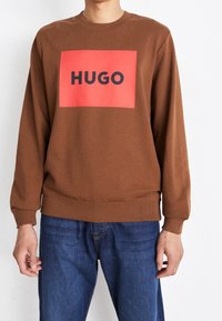 Sweatshirt marron avec un logo rectangulaire rouge affichant "HUGO" en noir. Comprend des poignets et un ourlet côtelés, coupe décontractée, texture douce.