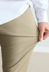 Beige trousers with a textured finish, pulled slightly from the waist, paired with a white collared shirt. Flat fabric, tailored fit.
