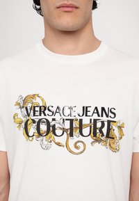 Balta medvilninė marškinė su juodu ir auksiniu puošniu raštu, kuriame yra tekstas "Versace Jeans Couture" ryškiu šriftu. Trumpomis rankovėmis ir apvalia kaklo iškirpte.