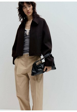 Mujer con una chaqueta marrón oscuro, camiseta gris, pantalones beige de pierna ancha, sosteniendo un bolso de piel negro contra un fondo liso y claro.