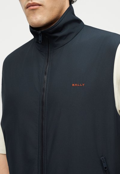 Άνδρας με μαύρο γιλέκο με φερμουάρ πάνω από λευκό κοντομάνικο πουκάμισο, με κόκκινο λογότυπο "BALLY" στο αριστερό στήθος και τσέπη με φερμουάρ στο πλάι.